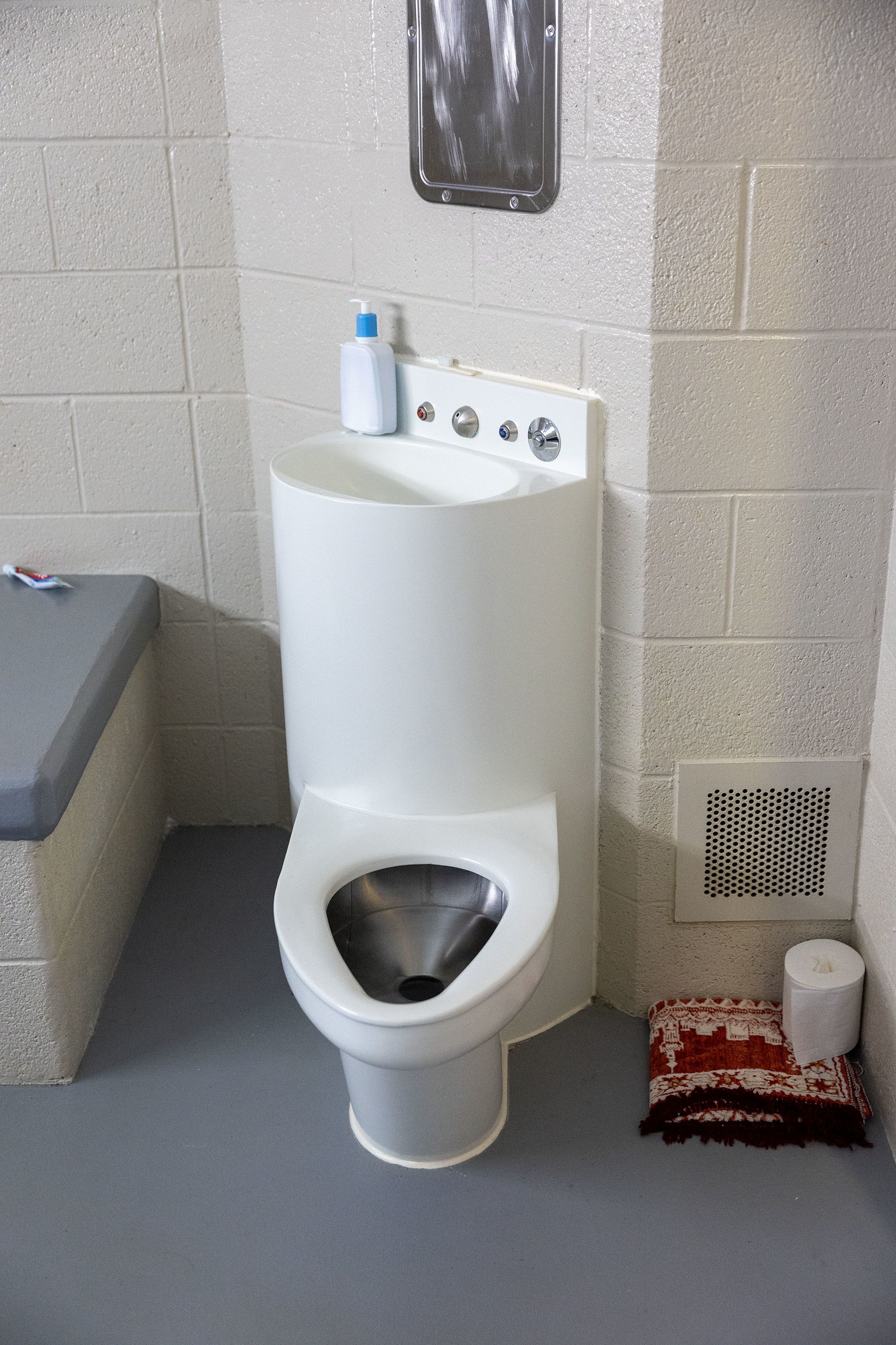 Basic facilities include a combination toilet and sink next to a bench and a folded-up prayer mat.  inside a cell at Decker Lake Youth Center.