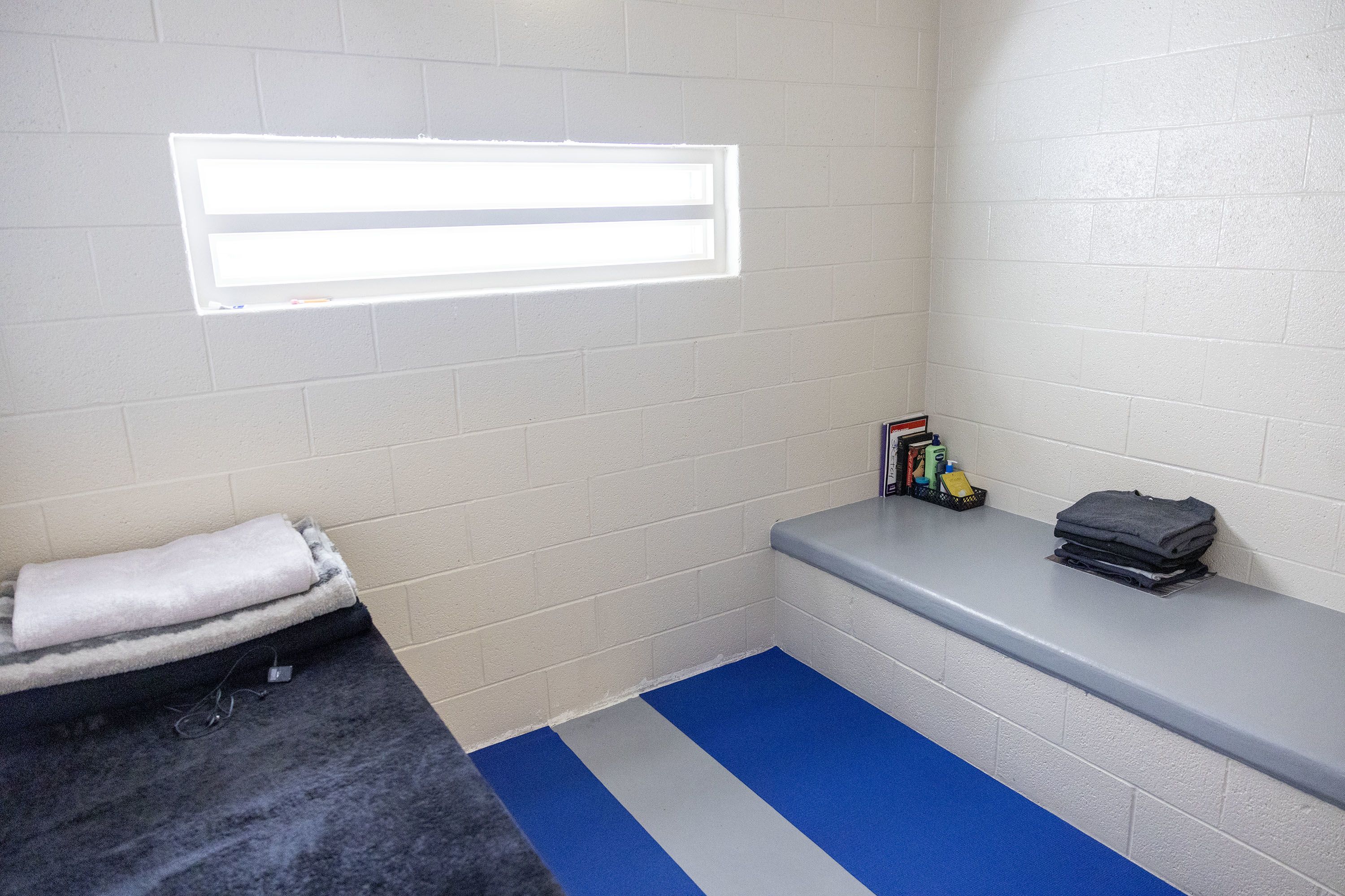 A look inside a cell at Decker Lake Youth Center, with a small bed, a bench, a few books, and a small window.