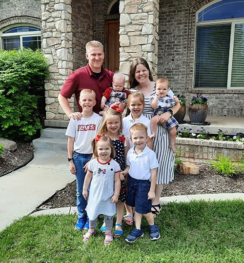 Sarah, a woman with long blonde hair in a dress, stands in front of her house with her husband and 7 children.