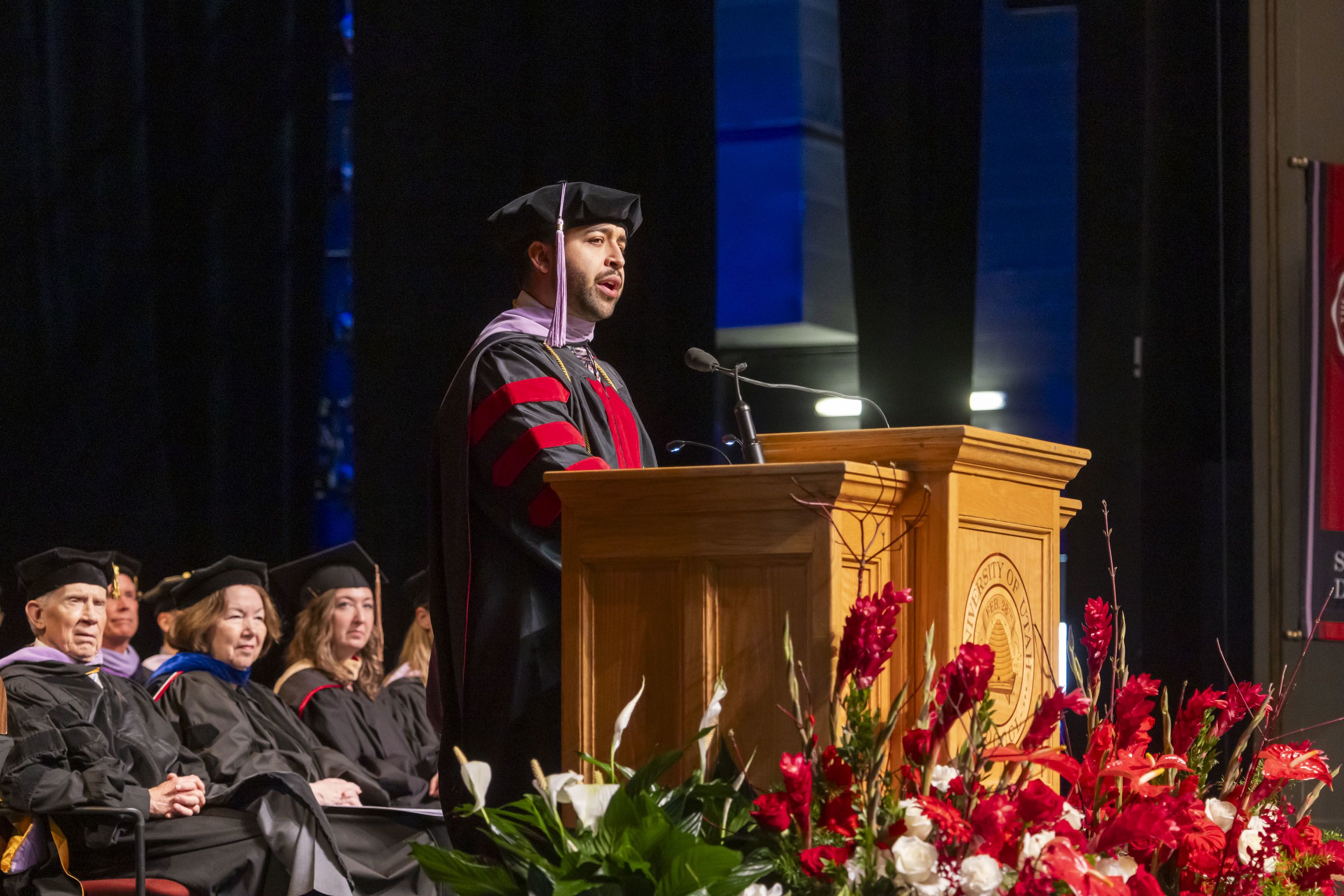 In beret and red stripped gown, Alex Rojas giving his commencement speech to his fellow 2024 graduates from the Kingsbury Hall stage as faculty look on. 